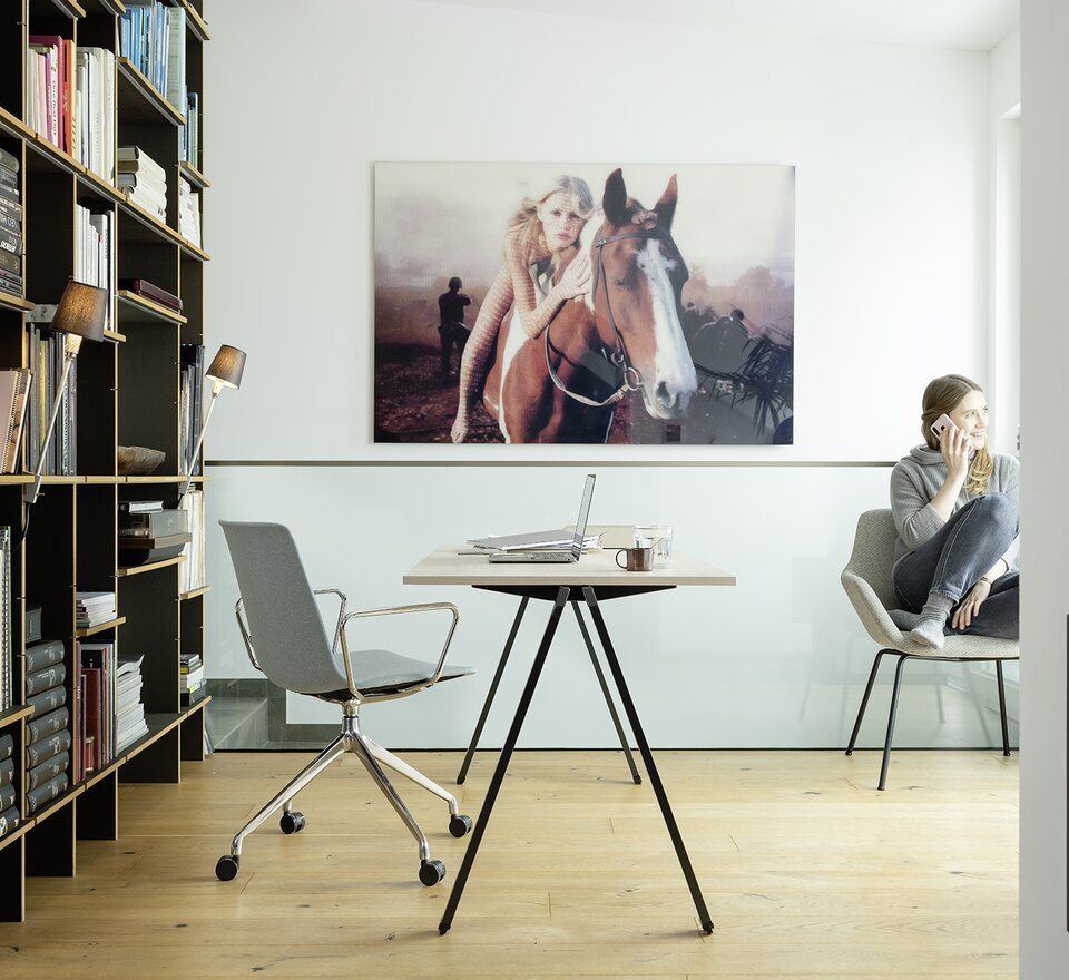 Desk with swivel chair. Woman is sitting behind it at the window and on the phone.